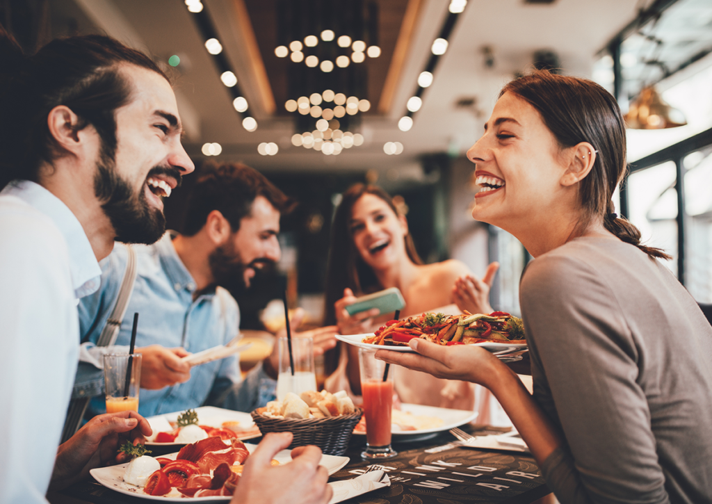 Group of happy people sitting at a restaurant table, smiling and laughing while enjoying their meal. The table is filled with delicious food and drinks, creating a warm and joyful atmosphere